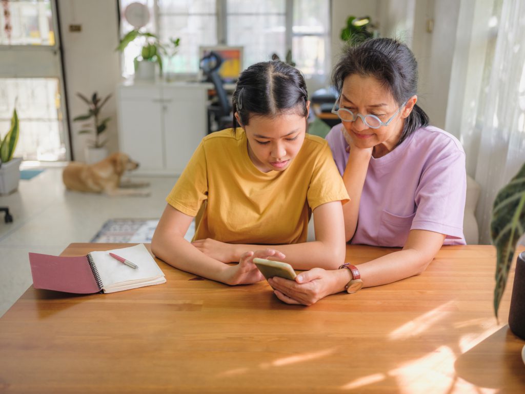 Happy Asian teenage daughter and mother using smart phone for online banking or video call with their dog lying down on the ground at home office